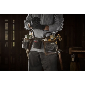 Почетна 54 A person wearing a DEWALT tool belt full of tools, including a measuring tape, screwdrivers, pencils, and a yellow and black cordless drill, standing in a workshop and putting on black work gloves.