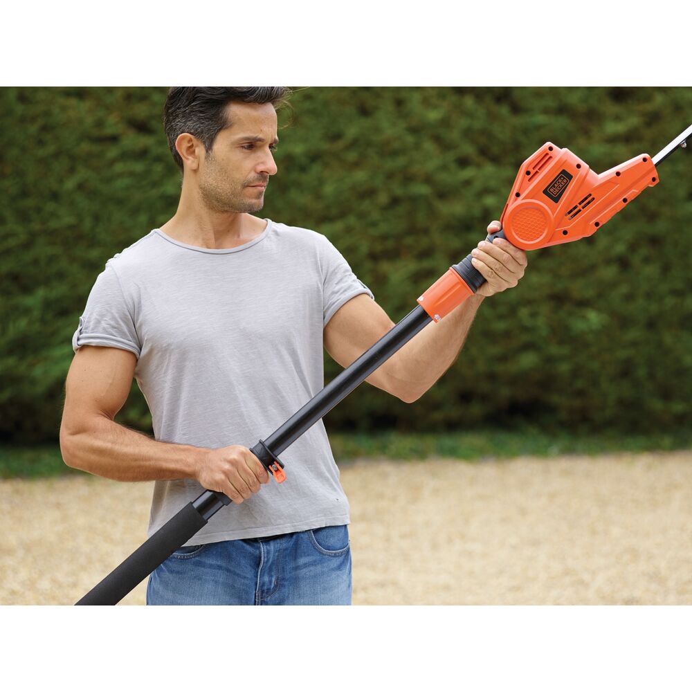 A person holding a BLACK+DECKER telescopic tool with an orange and black handle outdoors, likely used for gardening or yard work. The person's face is blurred.
